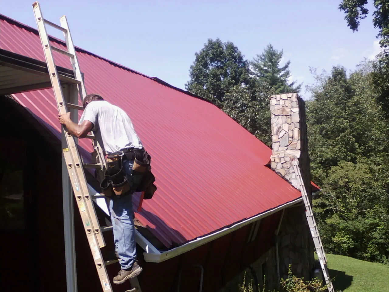 Roofer on a ladder beside a red metal roof and stone chimney — Soffit & Fascia Repair by Roofing Brothers in Highwood
